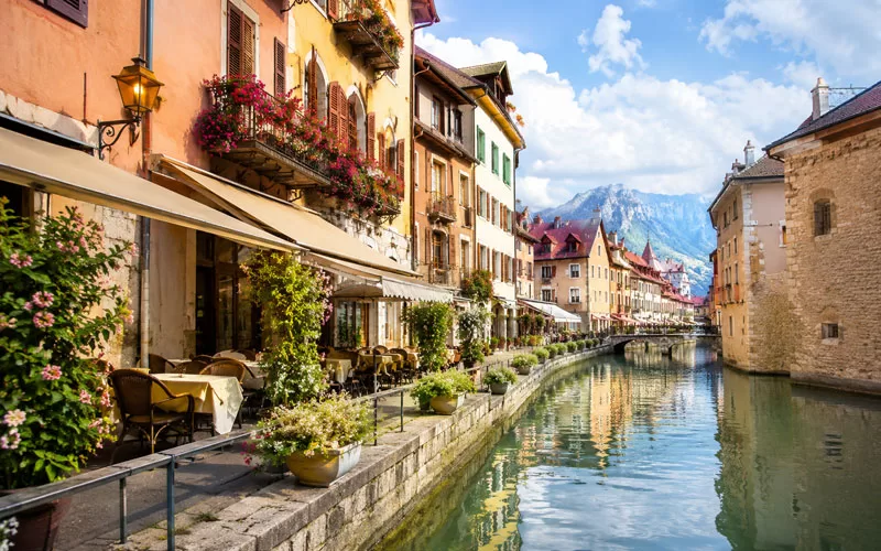 Colorful canal-side buildings with flower boxes, café seating, calm water, and mountain views in a charming European town