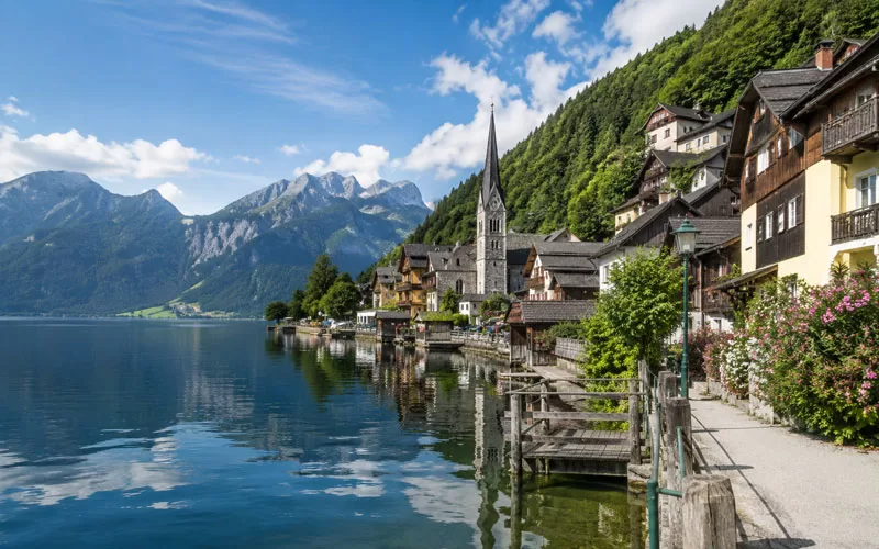 Peaceful lakeside village with mountain views, calm water reflections, waterfront homes, flowers, and a church steeple