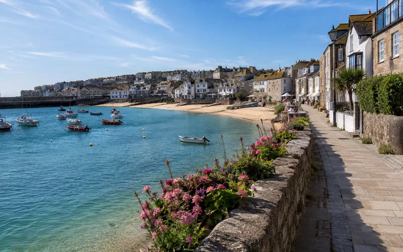 Coastal harbor town with blue water, small boats, sandy beach, seaside buildings, stone walkway, and pink flowers
