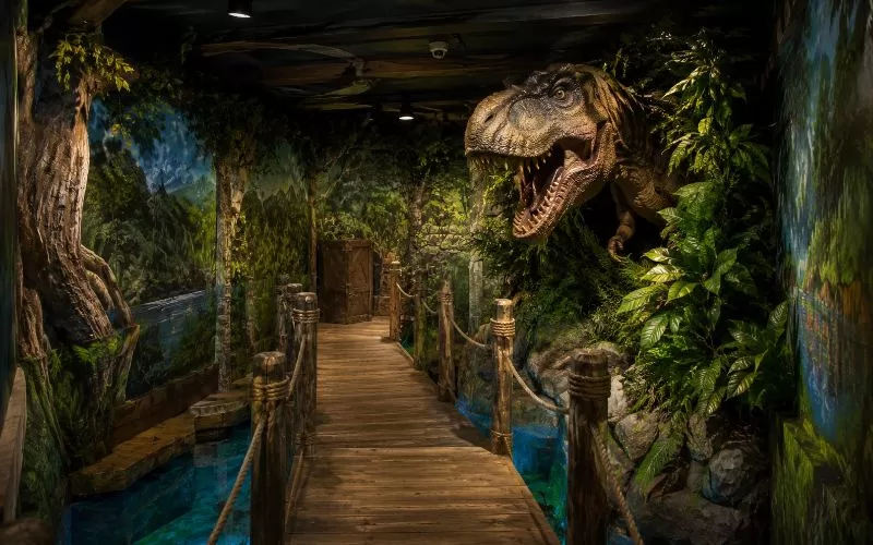 Jurassic-inspired escape room scene with a wooden plank bridge over fake water, painted jungle walls, tropical foliage, and a dinosaur emerging from the set.