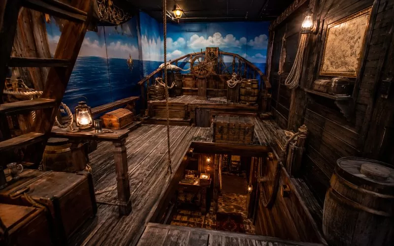 Pirate-inspired escape room scene with a wooden ship deck, ladder, hatch to a lower level, ropes, barrels, lanterns, treasure chests, and ocean-painted walls.