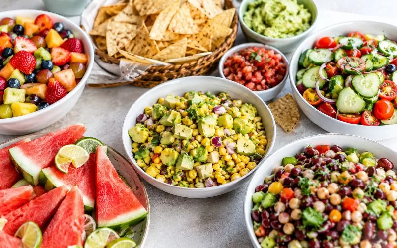 Fresh summer side dishes with watermelon, fruit salad, corn salad, cucumber tomato salad, bean salad, chips, salsa, and guacamole