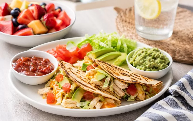 Light chicken tacos with salsa, guacamole, lettuce, avocado, and fruit on a bright table