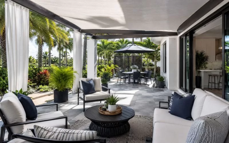 Shaded Florida patio with sliding glass doors, outdoor curtains, a pergola-style shade cover, navy and white seating, and palm trees.