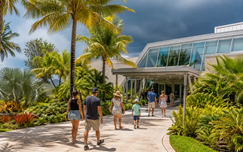 Visitors walk toward a modern indoor attraction surrounded by tropical landscaping as summer storm clouds gather.