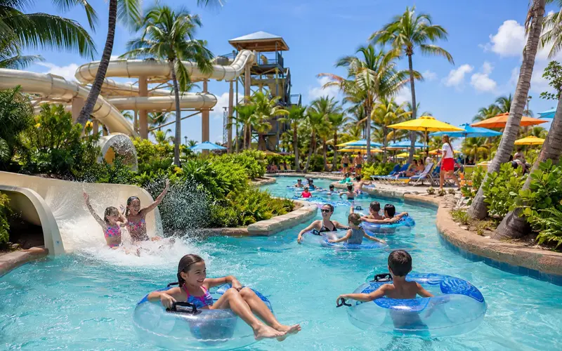 Families float along a tropical lazy river beside a curving waterslide, palm trees, and colorful umbrellas.